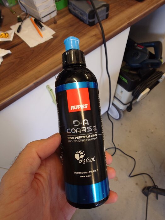 Flaska med Rupes D-A Coarse poleringsmedel hålls i en hand. Bakgrund med en arbetsbänk och verktyg.