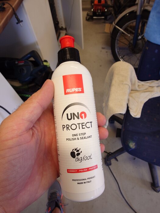 Rupes UNO Protect polishflaska i en hand, omgiven av garageutrustning.