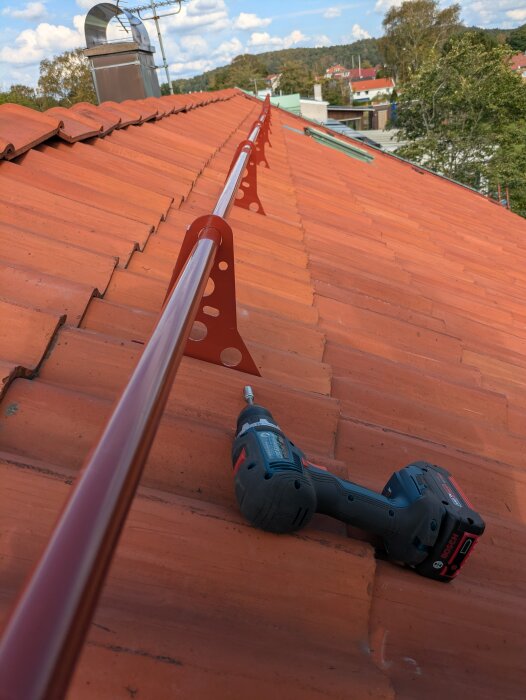 Tak med nyinstallerat nockräcke och skruvdragare på tegelpannor, omgivning med träd och hus i bakgrunden.