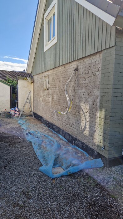 Fasad av gammalt lerstenshus från 1800-talet i Skåne med putsad gavel, sladdar hänger, blå presenning ligger vid marken.