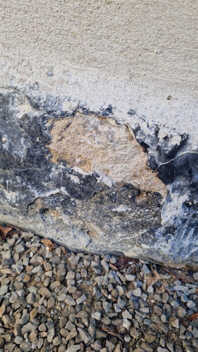 Skadat fundament på gammalt hus med exponerad tegelsten och sliten puts, omgivet av grus.