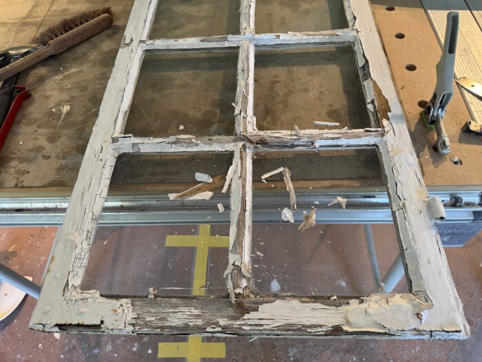 Slitet gammalt fönster med åtta glasrutor och flagnande färg, ligger på arbetsbänk för renovering. Fönsterbågens skick är dåligt.