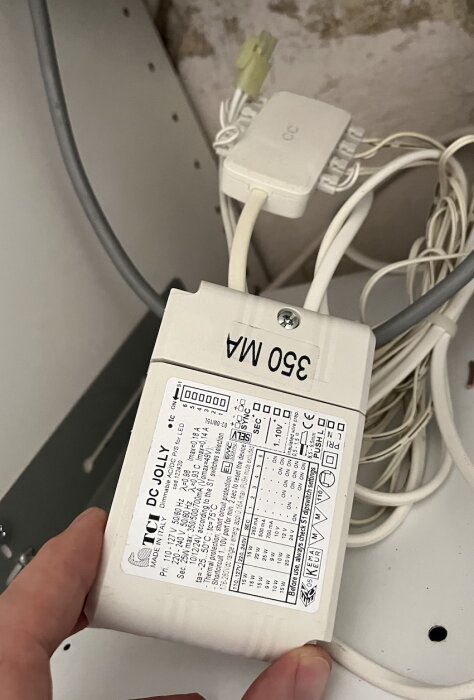 En Jolly-drivare märkt 350 mA, hållen i handen med anslutna kablar, mot en bakgrund av en vägg och andra elektriska kopplingar.