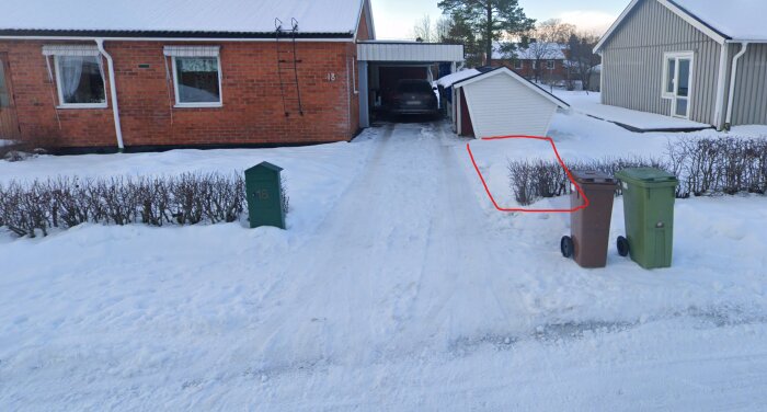 Uppfart med snö vid ett radhus, rabatter markerade med röd linje, två soptunnor till höger, bil i carport, snötäckt mark.