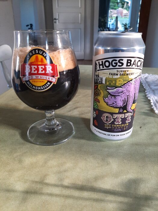 Ett glas mörk öl med skumkrona bredvid en burk med etikett "Hogs Back Brewery". Sokulär bakgrund inomhus.