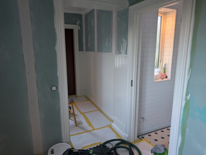 Rum under renovering med pärlspontspanel och blågröna väggar, där grundfärgen har tagit slut. Dörröppning in till kaklat badrum.