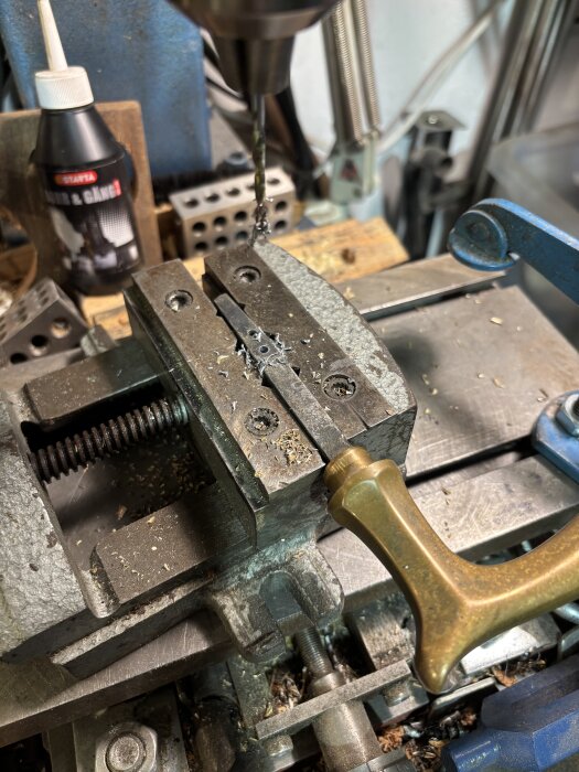 Borrmaskin borrar genom metallföremål i verkstad. Verktyg och oljeflaska syns i bakgrunden.