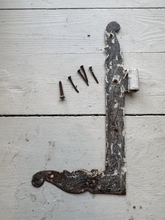 Rostigt gammalt gångjärn med färgrester, tillsammans med fyra gamla spikar och en modern skruv, på en vit yta redo för renovering.