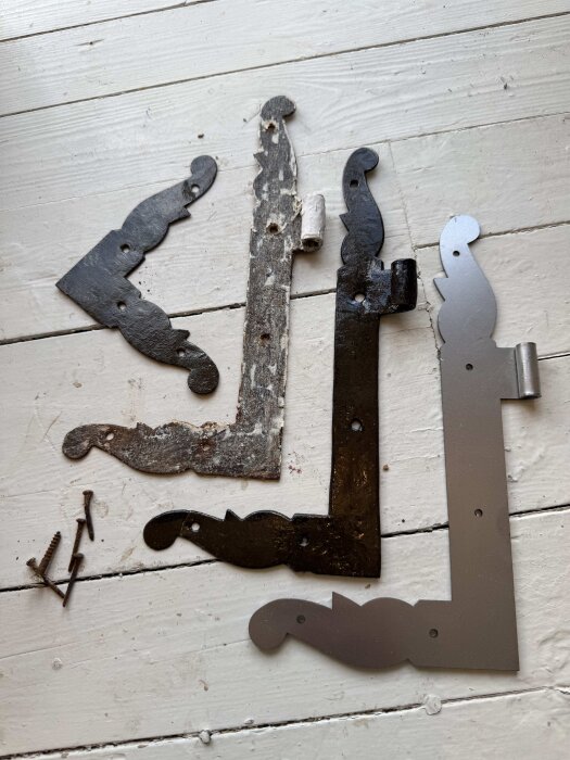 Gamla och nya gångjärn på ett vitt underlag. De är av olika skick, från rostiga till blanka och maskintillverkade. Flera skruvar ligger bredvid.