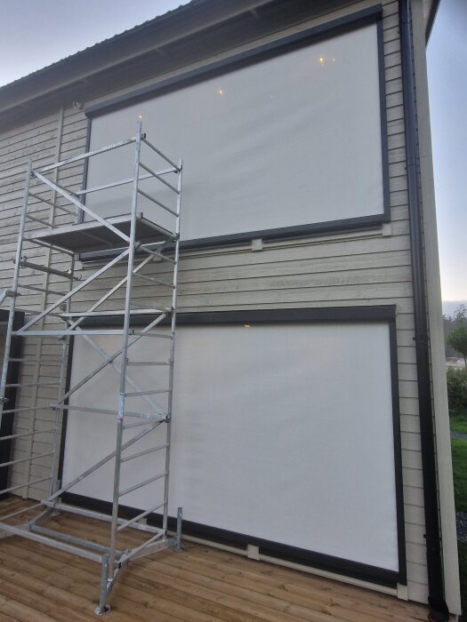 Två vita, breda markiser på en träfasad med ett byggställning vid sidan.