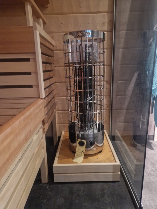 Bastuaggregat placerat på en hemmagjord träram för att skydda golvets matta från värme, med en träbänk och glasvägg i bakgrunden.