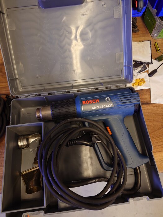 Blå Bosch varmluftspistol modell GHG 650 LCE med sladd i en öppen verktygslåda på ett träbord.