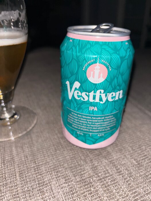 En ölburk av märket Vestfyen IPA står bredvid ett halvfullt ölglas på ett bord.