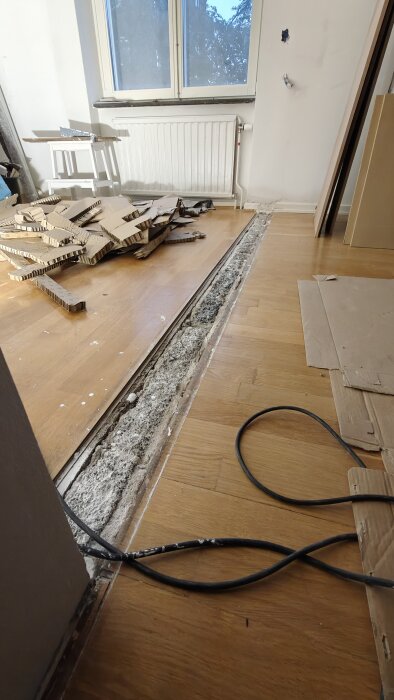 Oppussingsprosjekt med åpning mellom parkettgulv og betong, pappbiter og en kabel på gulvet, i et rom med vindu og radiator.