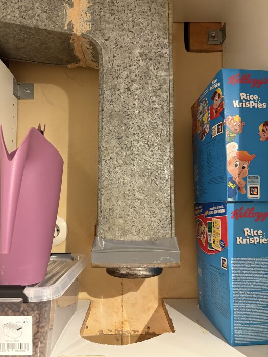 En köksventilationskanal i metall omgivet av kartonger och plastförvaring i ett skåp.