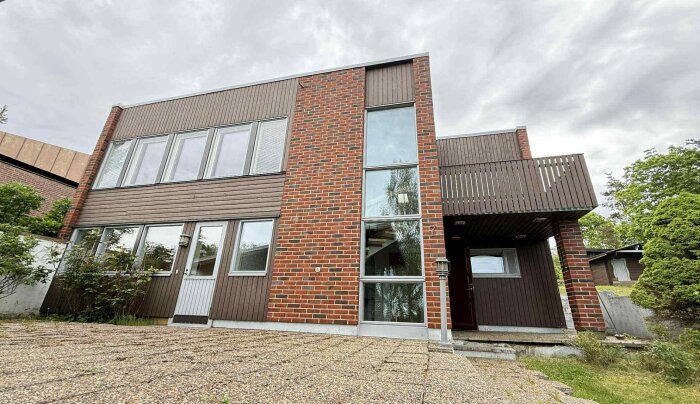 Rött tegelhus från 1974 med flera nya fönster och platt tak, fotat från framsidan med vy över entrén och omgivande trädgård.