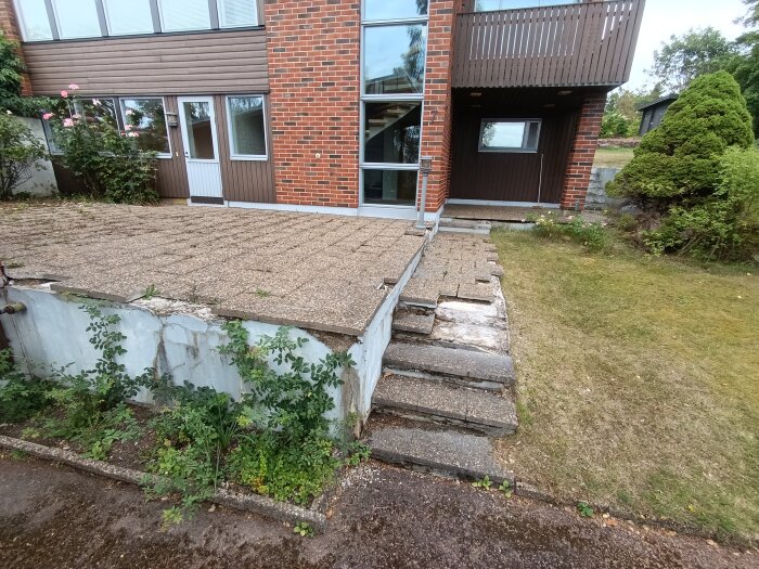 Uteplats med slitna och delvis trasiga plattor framför ett tegelklätt hus med dörr och fönster.