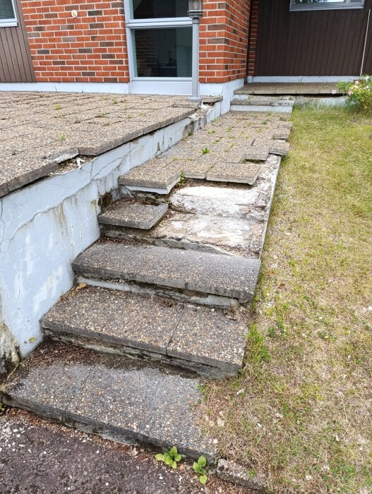 Slitna betongplattor på en trappa framför ett tegelhus, med gräs och växter vid sidan.