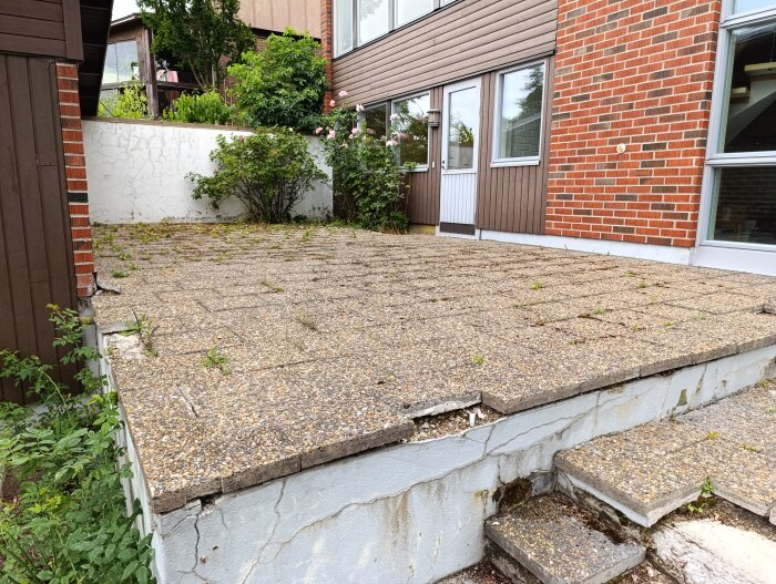 En uteplats med väderbitna plattor och spruckna kanter framför ett tegelhus.