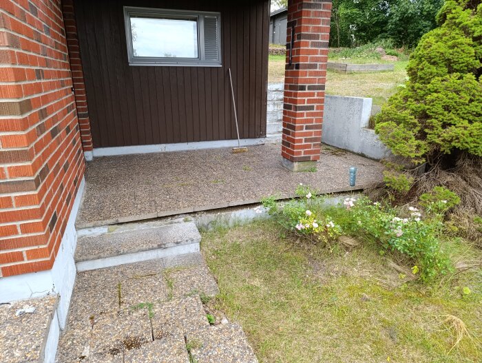 En uteplats med korniga betongplattor vid ett hus med röd tegelfasad, trappa i förgrunden, och blommande buskar vid sidan.