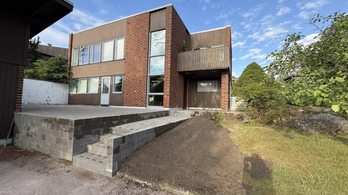 Byggnad med två våningar i tegel och träfasad, plattsatt uteplats med trappa, och nyplanterad jord framför trappan.