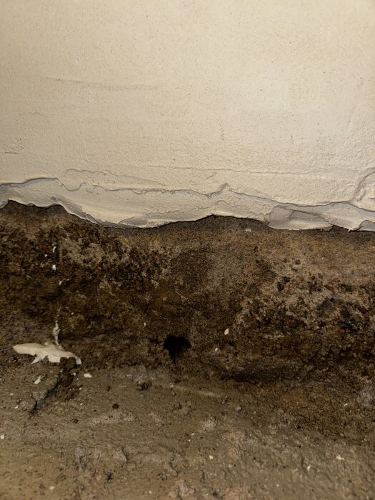 En närbild på en vägg med ett tydligt hål vid basen där vatten kan tränga in. Väggputsen är sprucken och marken är synlig.