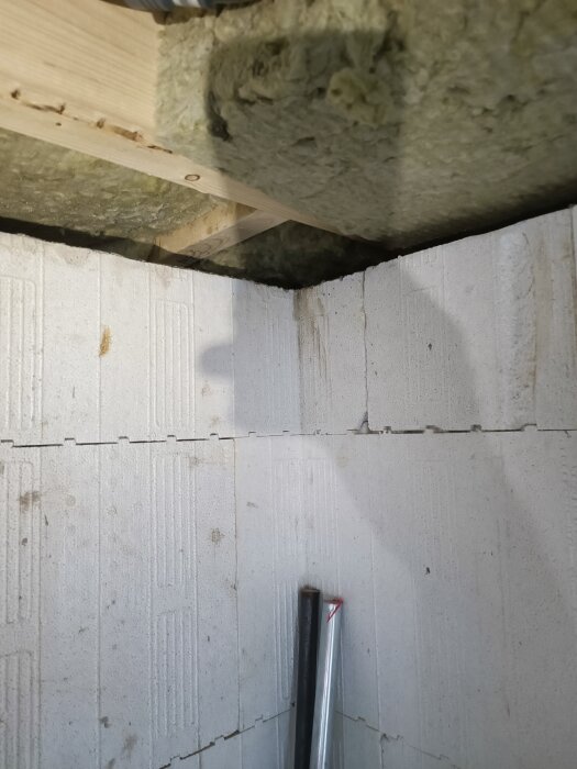 Källarhörn med thermomur, synligt bjälklag och isolering i taket, två rör står mot väggen, visar byggprojektets detaljer.