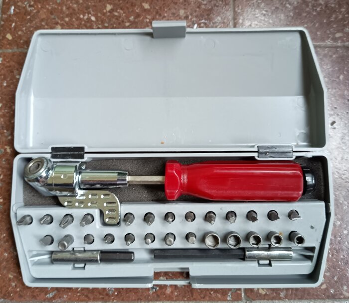 Vinkelskruvmejsel med rött handtag och olika bits i en öppen vit verktygsbox.