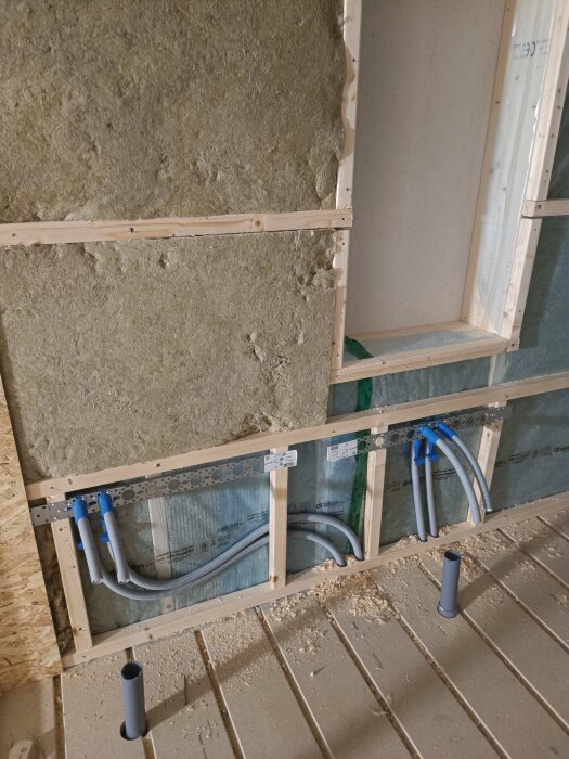 Isolering, träreglar och rör ovanpå spånskiva på en byggarbetsplats, med plast som kläms mot överkanten av skivan.