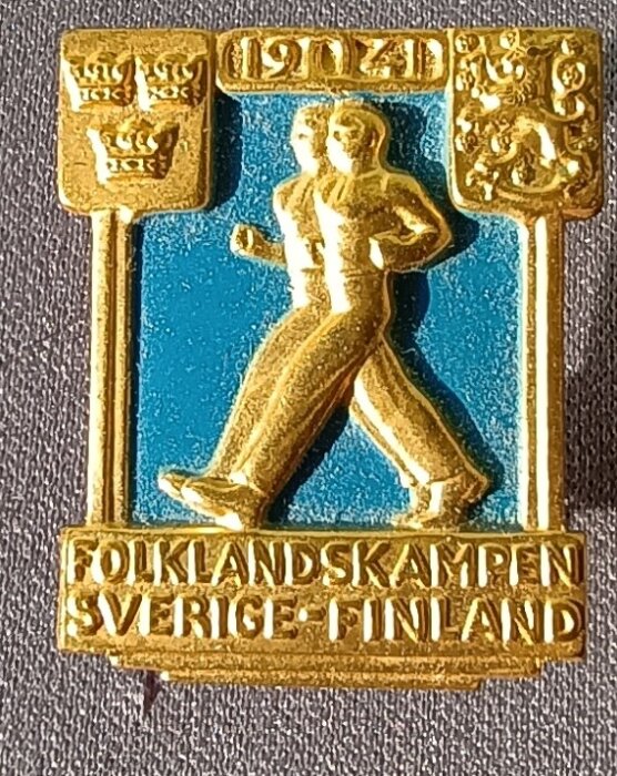 Märke från 1941 års Folklandskampen mellan Sverige och Finland med två gående figurer, svenska och finska vapen och texten "FOLKLANDSKAMPEN SVERIGE - FINLAND".
