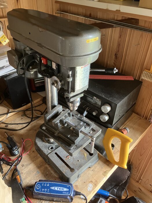 Borrmaskin på arbetsbänk i torp, med olika verktyg runtomkring, inklusive en såg och en laddare.