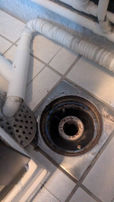 Golvbrunn i ett 70-talshus med avsaknad av vattenlås. Synliga rör och kaklat golv.