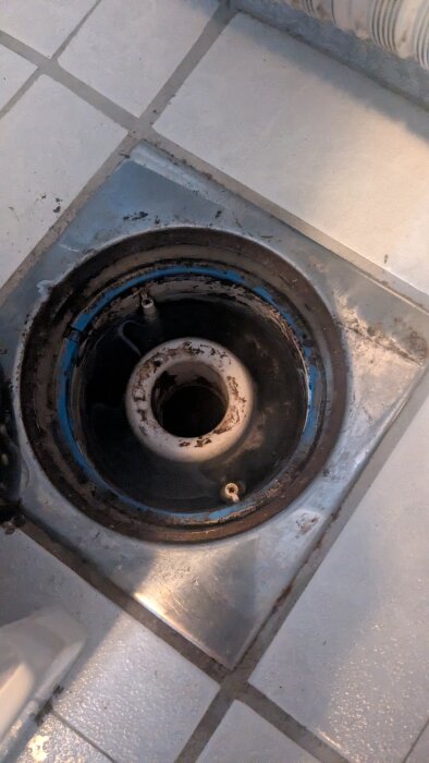Golvbrunn med öppet lock, omgiven av kakelgolv. Möjligen saknar vattenlås insats vilket leder till luktproblem.