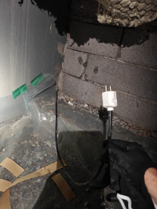 Elektrisk sladd och kontakt i krypgrund, hålls av en hand med handske. Underlaget består av makadam täckt av plastfilm.