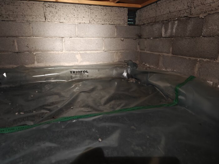 Krypgrund med makadam täckt av Trioplast-ångspärr samt tegelväggar och ventilationsöppning synlig i hörnet.