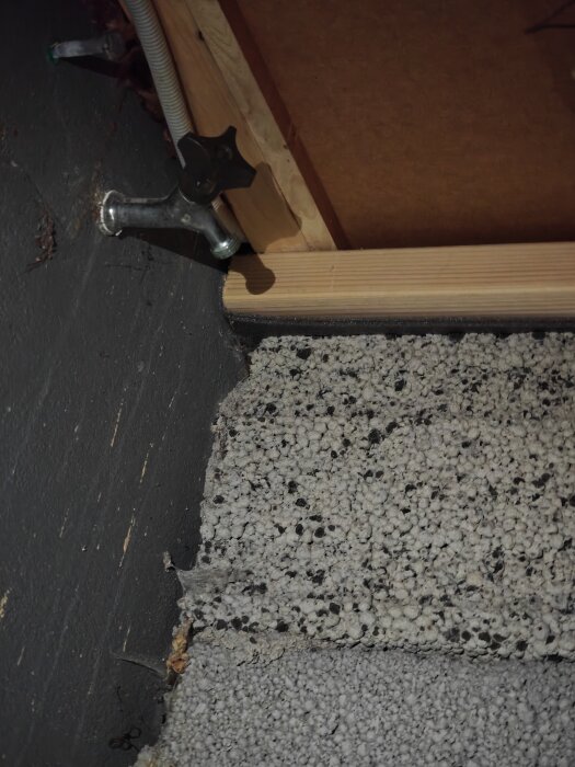 Vattenutkastare nära eluttag och ventilationskanal, med två svarta rör för golvvärme synliga bakom träpanel och grå betongvägg.