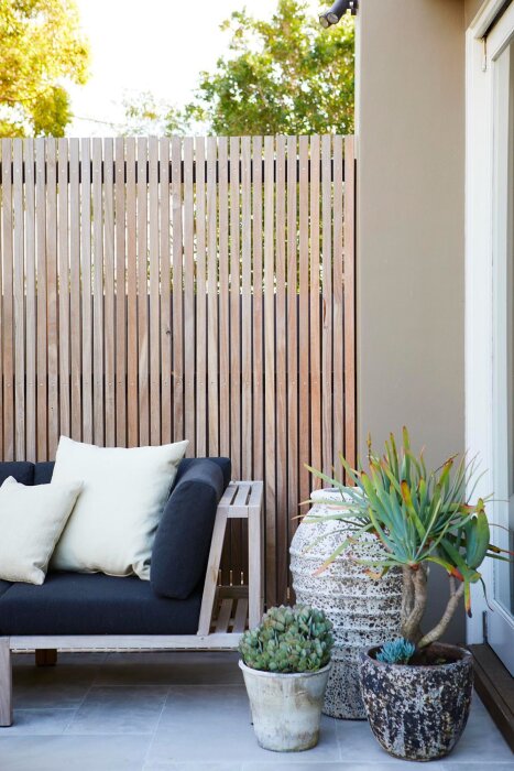 Vertikalt ribbat träplank på uteplats med svart soffa, vita kuddar och krukväxter i förgrunden.
