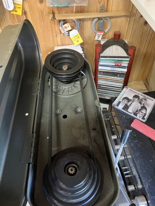 En maskin med öppen topp som visar en rem och remskivor monterade inuti, omgiven av verktyg och andra föremål i bakgrunden.