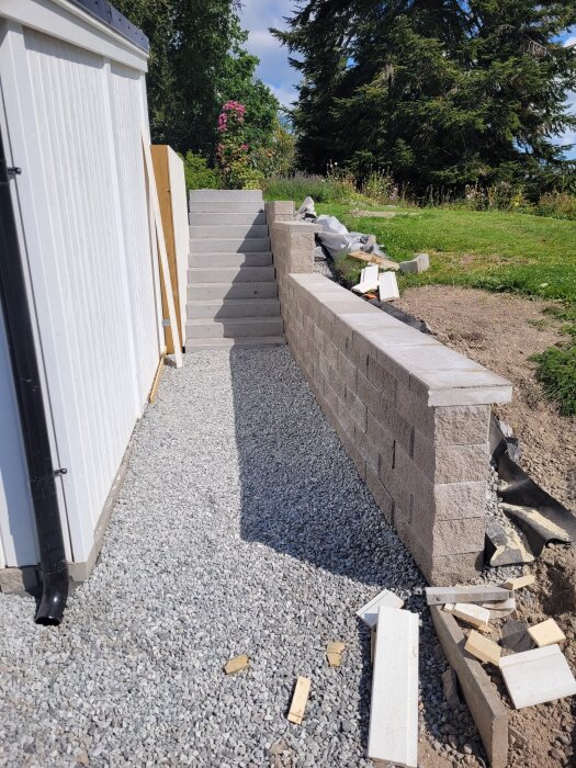 Grusgång med intilliggande stenmur och trappa vid vit byggnad, omgiven av grönska.