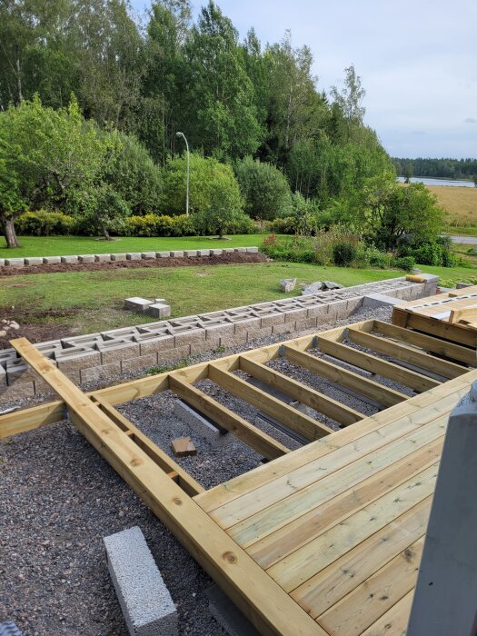 En pågående träkonstruktion med en mur av betongblock i en lummig trädgårdsmiljö.