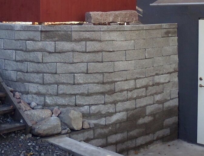 Stenmur med grå block, 2 meter hög, byggd med geonät och bergkross. Stenarna är staplade med små dekorationer ovanpå och vid sidan.