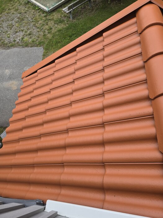 Nymålat tak av farstukvist med tegelliknande pannor i rödbrun nyans, sett från ovan. Gräsmatta och stenlagd yta syns i bakgrunden.