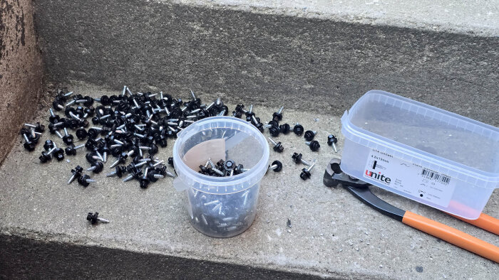 Avklippta svart- och silverfärgade skruvar på betongsteg, bredvid en plastbehållare och en avbitartång med orange handtag.