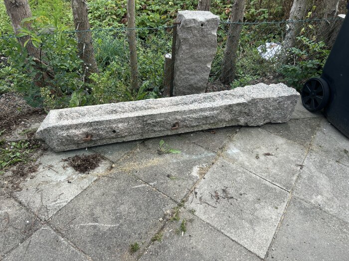 Grindstolpe av sten liggandes på marken, omgiven av plattor och stängsel, i en trädgård med buskar och träd i bakgrunden.