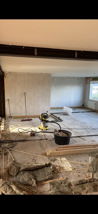 Construction room with removed floor components, exposed pipes, insulation, vacuum, and tools scattered. Room under renovation for underfloor heating installation.