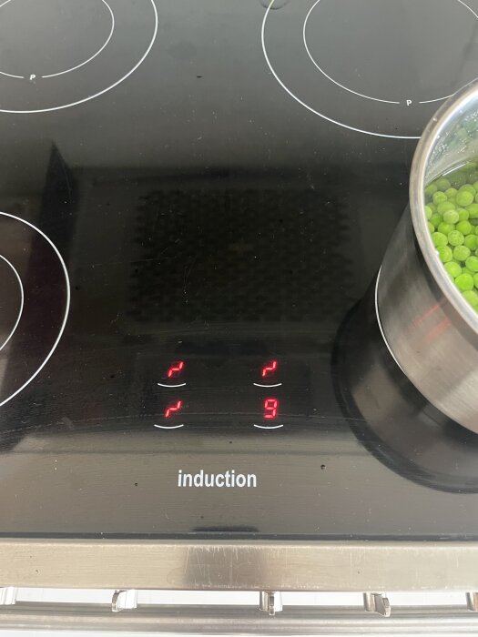 Induktionshäll med blinkande symboler och siffror, pott med gröna ärtor på en av kokzonerna.