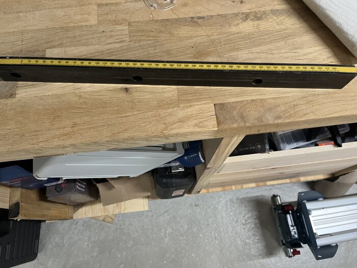 Mätstång med gul skala ligger på ett träbord, omgiven av verktyg och tillbehör i en verkstadsmiljö.