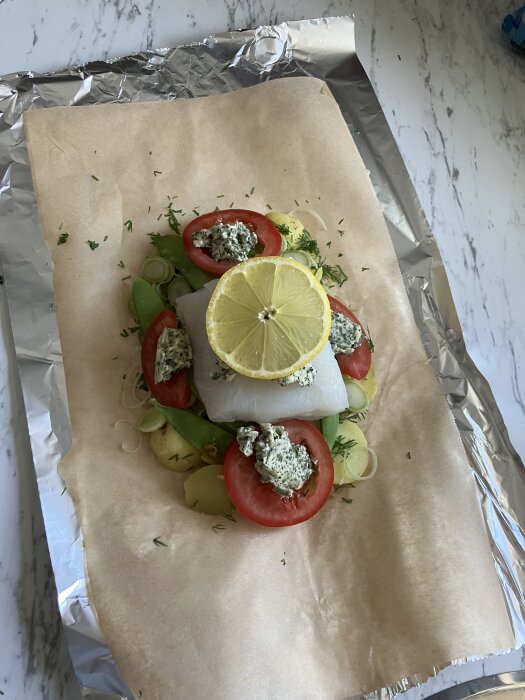 Fiskpaket med torsk, potatis, fänkål, tomat, purjolök och sockerärtor, toppat med örtsmör och citronskiva på bakplåtspapper.