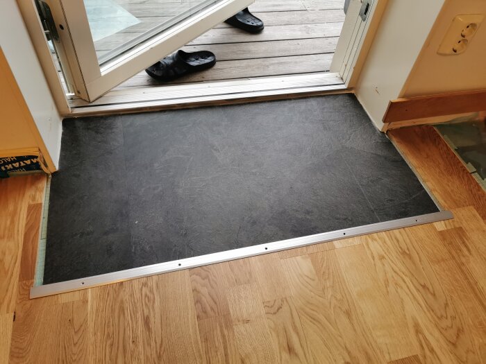 Ingång med öppen dörr, svart vinylgolv omslutet av träparkett. Lerskor utanför. Klass 33 vinyl och XPS skivor används som underlag.