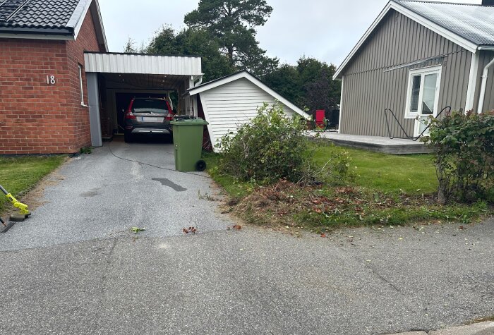 Uppfart med bil, klippt häck vid tomtgräns, gröna soptunnor och två hus, ett i tegel och ett i trä.